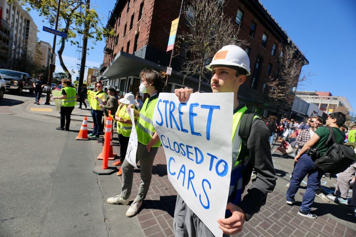 A person in a hard hat and yellow safety vest holds a sign reading "street closed to cars" while standing at the intersection of Telegraph and Durant avenues during a rally near UC Berkeley.